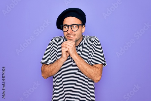 Handsome man with blue eyes wearing striped t-shirt and french beret over purple background laughing nervous and excited with hands on chin looking to the side