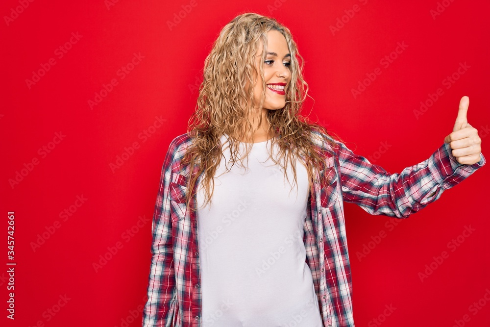 Fototapeta premium Young beautiful blonde woman wearing casual shirt standing over isolated red background Looking proud, smiling doing thumbs up gesture to the side