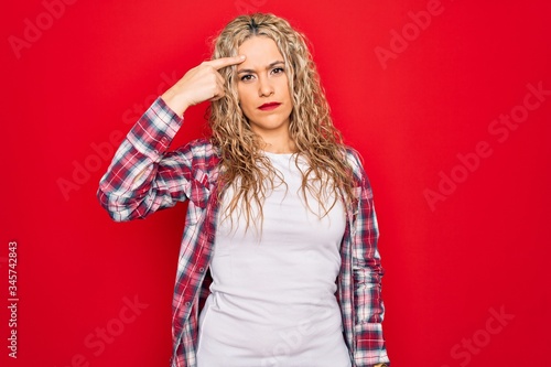 Young beautiful blonde woman wearing casual shirt standing over isolated red background pointing unhappy to pimple on forehead, ugly infection of blackhead. Acne and skin problem