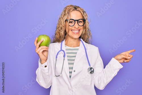 Young beautiful blonde doctor woman wearing stethoscope holding green apple fruit Smiling happy pointing with hand and finger to the side