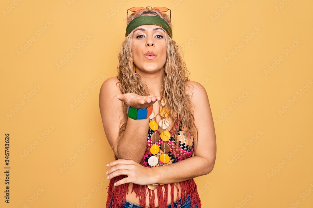 Beautiful blonde hippie woman wearing sunglasses and accessories over yellow background looking at the camera blowing a kiss with hand on air being lovely and sexy. Love expression.