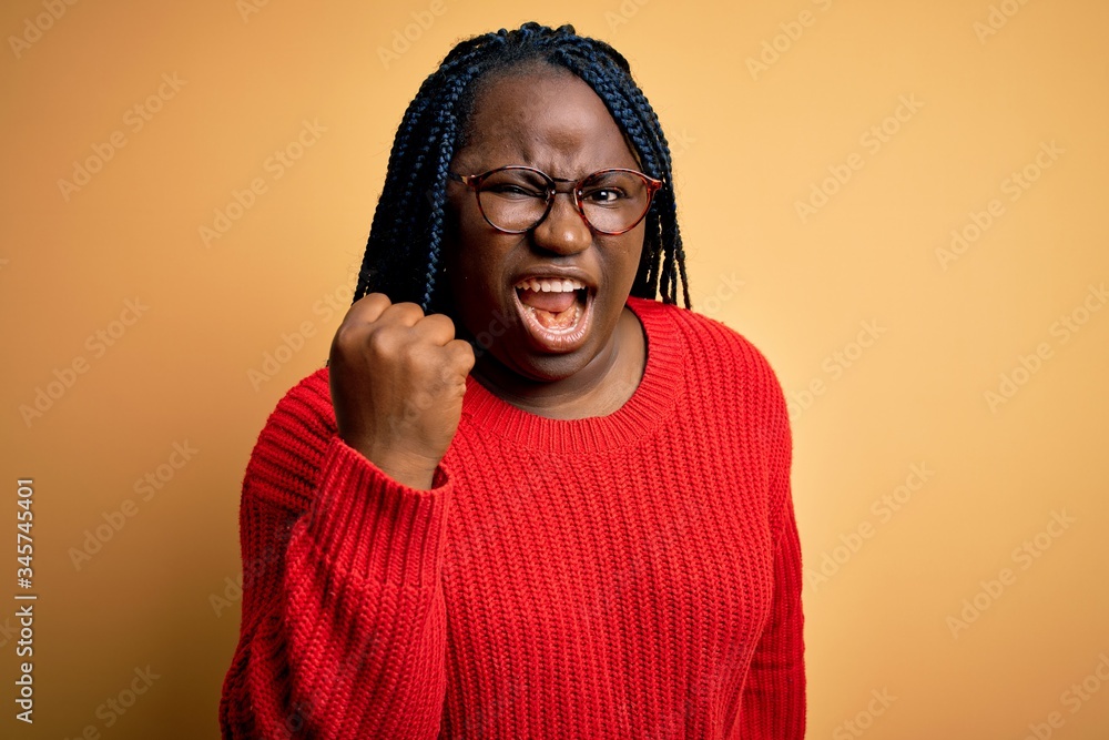 African american plus size woman with braids wearing casual sweater over yellow background angry and mad raising fist frustrated and furious while shouting with anger. Rage and aggressive concept. African american plus size woman with braids wearing casual sweater over yellow background angry and mad raising fist frustrated and furious while shouting with anger. Rage and aggressive concept.