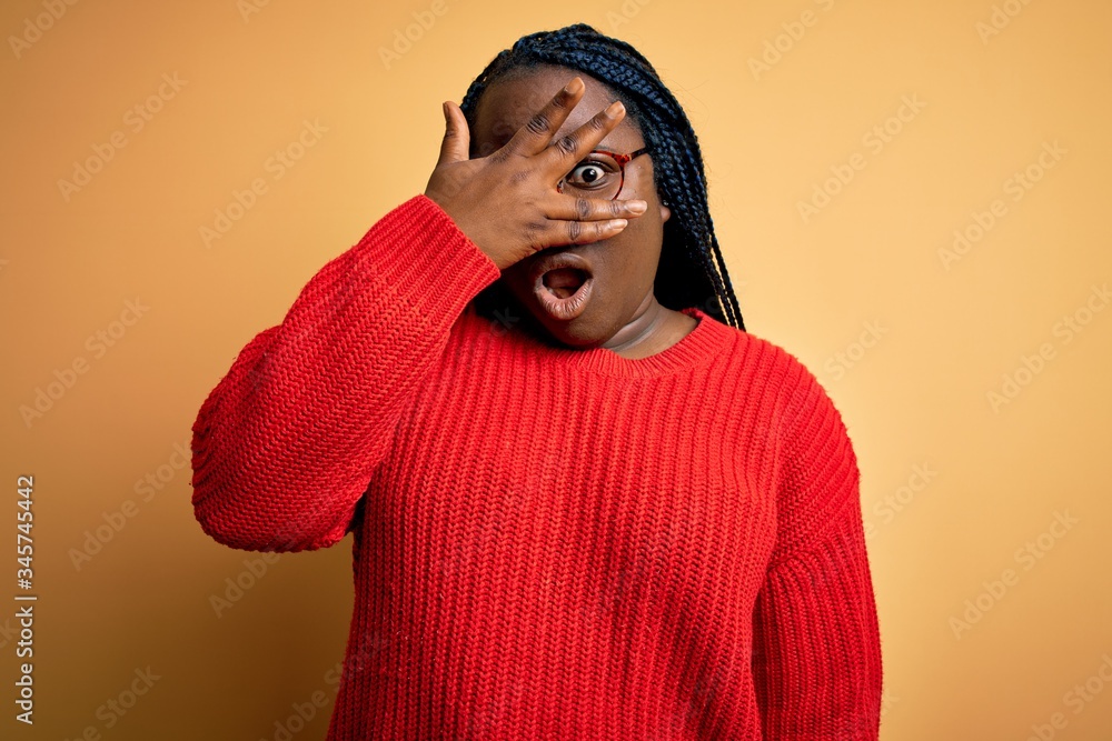 African american plus size woman with braids wearing casual sweater over yellow background peeking in shock covering face and eyes with hand, looking through fingers with embarrassed expression. African american plus size woman with braids wearing casual sweater over yellow background peeking in shock covering face and eyes with hand, looking through fingers with embarrassed expression.