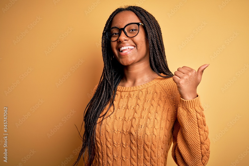 Young african american smart woman wearing glasses and casual sweater over yellow background smiling with happy face looking and pointing to the side with thumb up. Young african american smart woman wearing glasses and casual sweater over yellow background smiling with happy face looking and pointing to the side with thumb up.