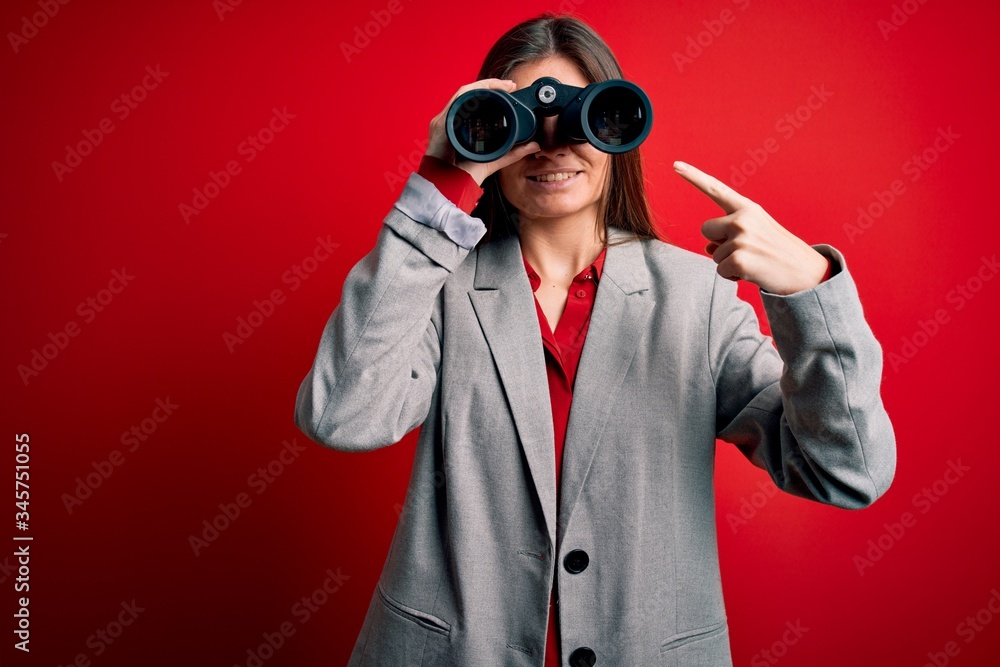 Young beautiful woman with blue eyes using binoculars over isolated red background very happy pointing with hand and finger Young beautiful woman with blue eyes using binoculars over isolated red background very happy pointing with hand and finger