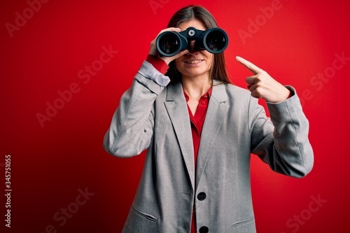 Young beautiful woman with blue eyes using binoculars over isolated red background very happy pointing with hand and finger