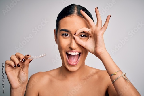 Young beautiful brunette woman holding dental aligner orthodontic to teeth corretion with happy face smiling doing ok sign with hand on eye looking through fingers photo