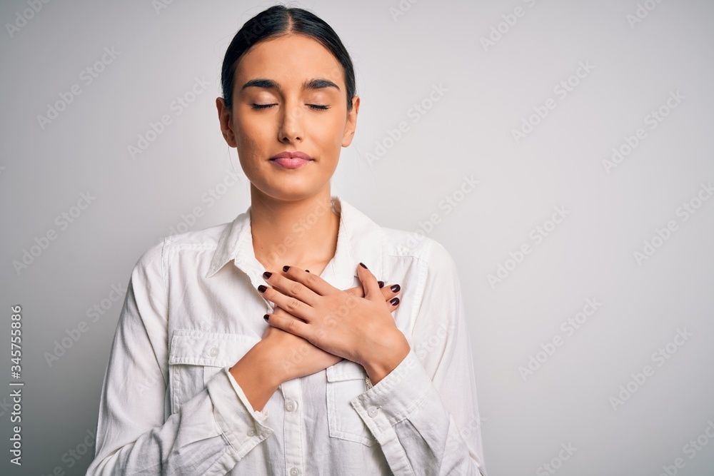 Young beautiful brunette woman wearing casual shirt over isolated white background smiling with hands on chest with closed eyes and grateful gesture on face. Health concept. Young beautiful brunette woman wearing casual shirt over isolated white background smiling with hands on chest with closed eyes and grateful gesture on face. Health concept.