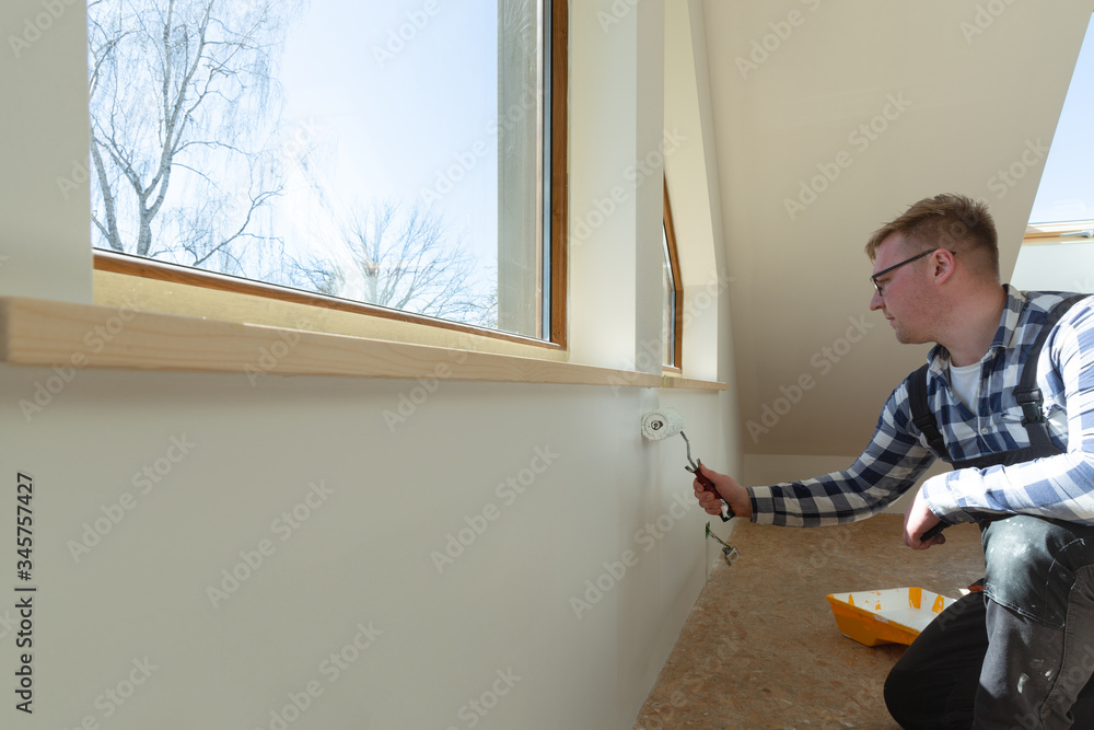 Home improvement concept, handyman painting a wall with a white paint near roof window in attic