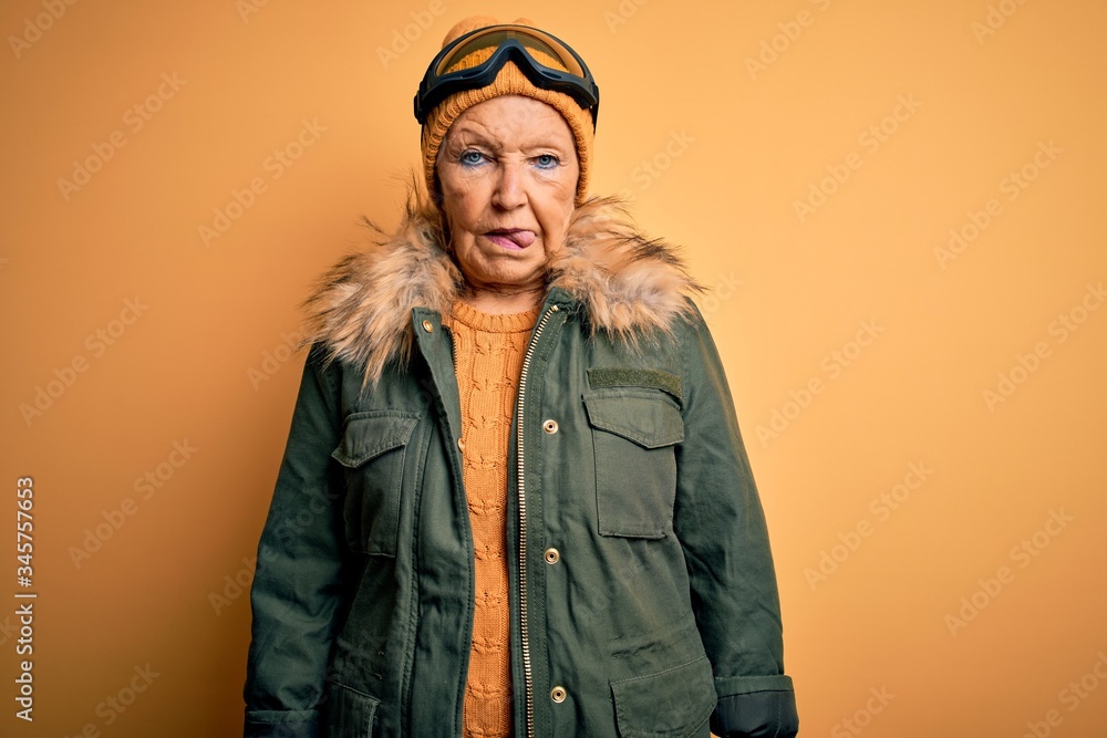 Senior beautiful grey-haired skier woman wearing snow sportswear and ski goggles sticking tongue out happy with funny expression. Emotion concept.