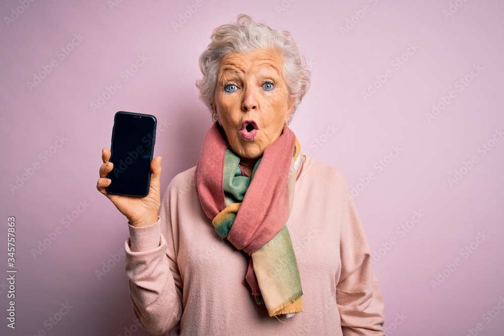 Senior beautiful grey-haired woman holding smartphone showing screen over pink background scared in shock with a surprise face, afraid and excited with fear expression Senior beautiful grey-haired woman holding smartphone showing screen over pink background scared in shock with a surprise face, afraid and excited with fear expression