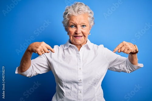 Senior beautiful woman wearing elegant shirt standing over isolated blue background looking confident with smile on face, pointing oneself with fingers proud and happy.