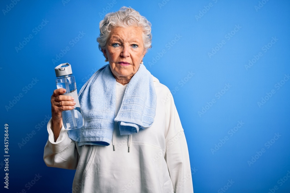 Senior beautiful sporty woman holding bottle of water standing over isolated blue background with a confident expression on smart face thinking serious Senior beautiful sporty woman holding bottle of water standing over isolated blue background with a confident expression on smart face thinking serious