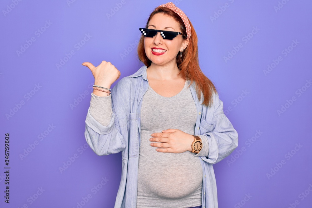 Fototapeta premium Young redhead pregnant woman expecting baby wearing funny thug life sunglasses smiling with happy face looking and pointing to the side with thumb up.