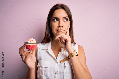 Young beautiful brunette woman eating chocolate cupcake over isolated pink background serious face thinking about question, very confused idea