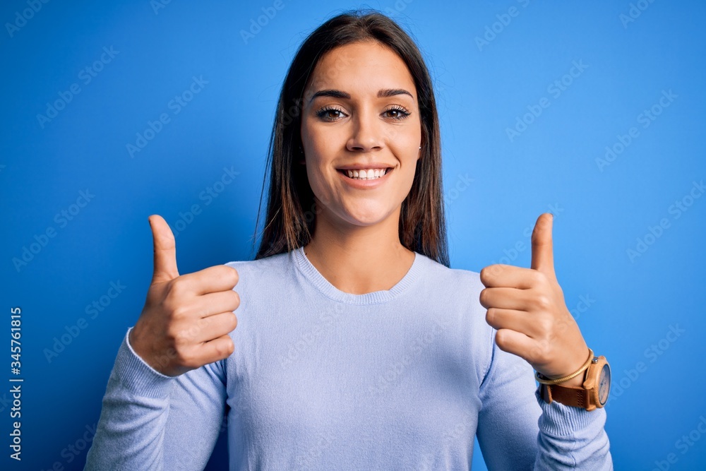 Young beautiful brunette woman wearing casual sweater standing over blue background success sign doing positive gesture with hand, thumbs up smiling and happy. Cheerful expression and winner gesture. Young beautiful brunette woman wearing casual sweater standing over blue background success sign doing positive gesture with hand, thumbs up smiling and happy. Cheerful expression and winner gesture.