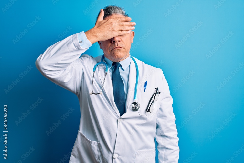 Middle age handsome grey-haired doctor man wearing coat and blue stethoscope covering eyes with hand, looking serious and sad. Sightless, hiding and rejection concept Middle age handsome grey-haired doctor man wearing coat and blue stethoscope covering eyes with hand, looking serious and sad. Sightless, hiding and rejection concept