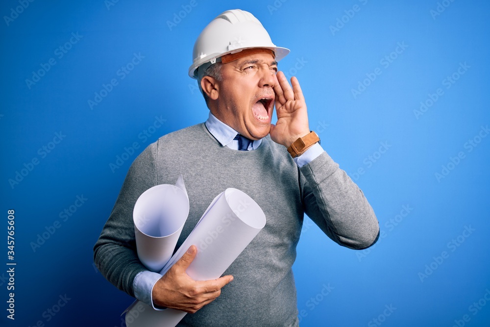 Middle age handsome grey-haired architect man wearing safety helmet holding blueprints shouting and screaming loud to side with hand on mouth. Communication concept. Middle age handsome grey-haired architect man wearing safety helmet holding blueprints shouting and screaming loud to side with hand on mouth. Communication concept.