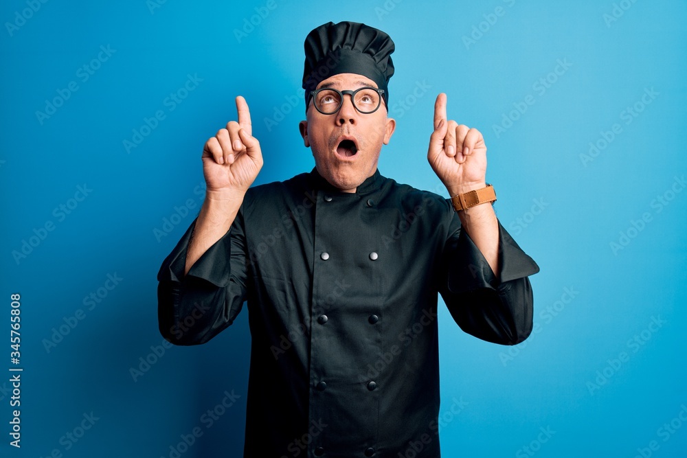 Middle age handsome grey-haired chef man wearing cooker uniform and hat amazed and surprised looking up and pointing with fingers and raised arms. Middle age handsome grey-haired chef man wearing cooker uniform and hat amazed and surprised looking up and pointing with fingers and raised arms.