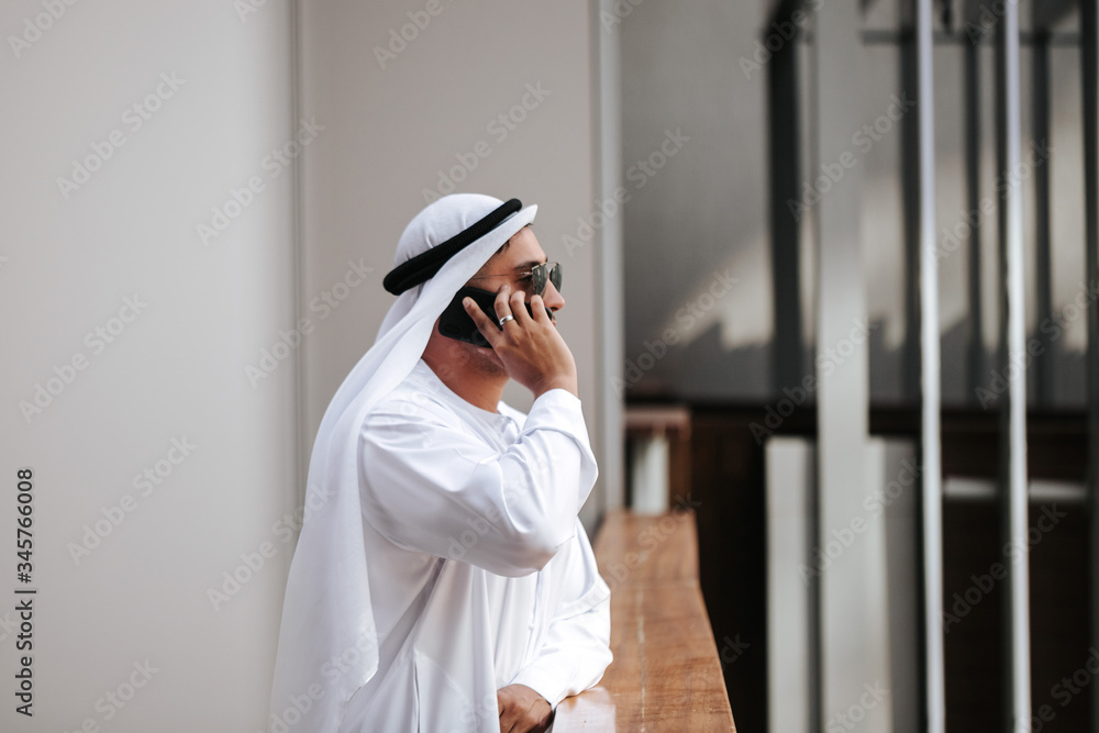 Emirati guy wearing traditional kandura in urban city emirates lifestyle talking to smart phone. Side view photography concept for Arab magazine.