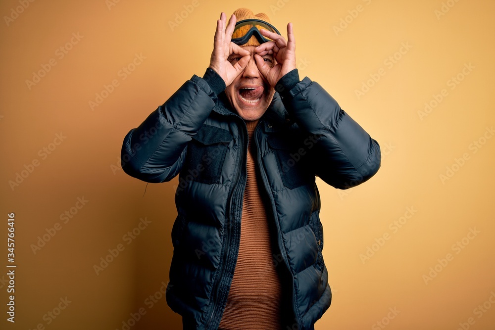 Middle age handsome grey-haired skier man on vacation wearing ski goggles doing ok gesture like binoculars sticking tongue out, eyes looking through fingers. Crazy expression. Middle age handsome grey-haired skier man on vacation wearing ski goggles doing ok gesture like binoculars sticking tongue out, eyes looking through fingers. Crazy expression.