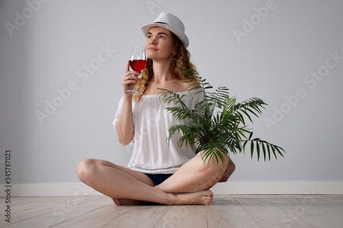 Girl with naked shoulders in white shirt and jeans shorts with glass manhattan cocktail and palm tree in hand sit on ceramic wooden tile floor nostalgia