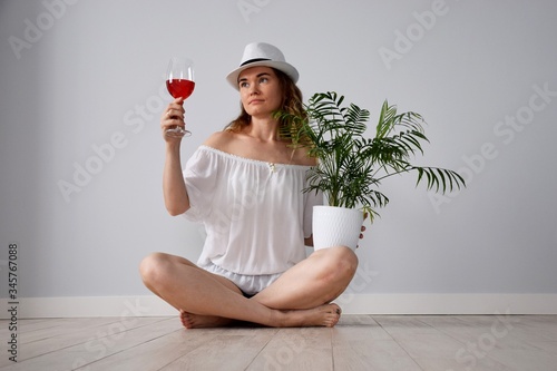 Girl with naked shoulders in white shirt and jeans shorts with glass manhattan cocktail and palm tree in hand sit on ceramic wooden tile floor nostalgia