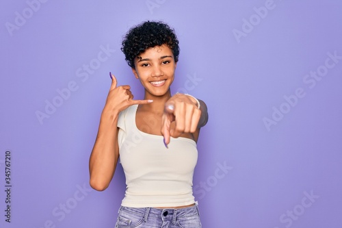 Young beautiful african american afro woman wearing casual t-shirt over purple background smiling doing talking on the telephone gesture and pointing to you. Call me.