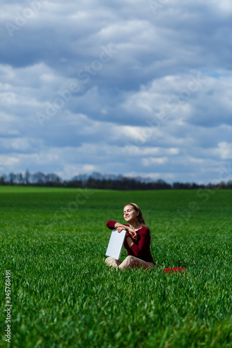 Young successful woman is sitting on green grass with a laptop in her hands. Rest after a good working day. Work on the nature. Student girl working in a secluded place. Workplace in nature © Дмитрий Ткачук
