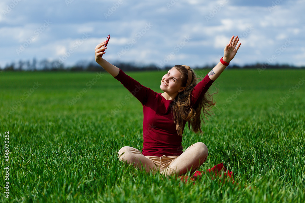 Young beautiful woman student with a phone in her hands sitting on the grass. Girl takes selfies and takes selfie pictures. She smiles and enjoys a warm day. Concept photo on smartphone Young beautiful woman student with a phone in her hands sitting on the grass. Girl takes selfies and takes selfie pictures. She smiles and enjoys a warm day. Concept photo on smartphone