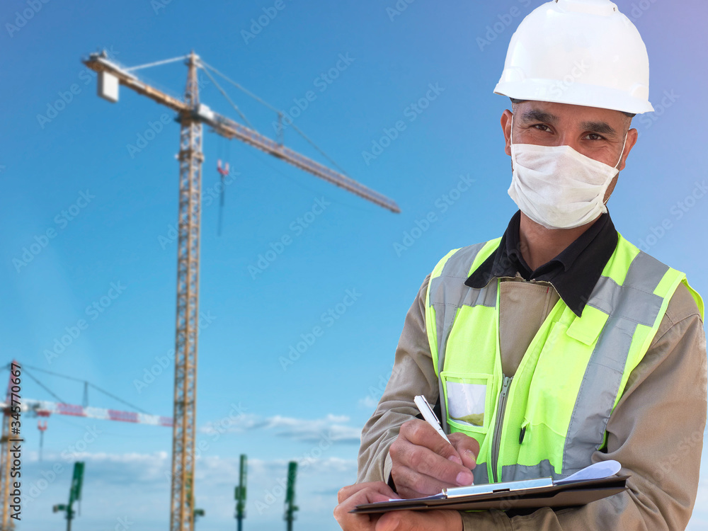 Construction worker in medical mask and white hardhat stands with clipboard opposite construction site with tower crane at background Construction worker in medical mask and white hardhat stands with clipboard opposite construction site with tower crane at background