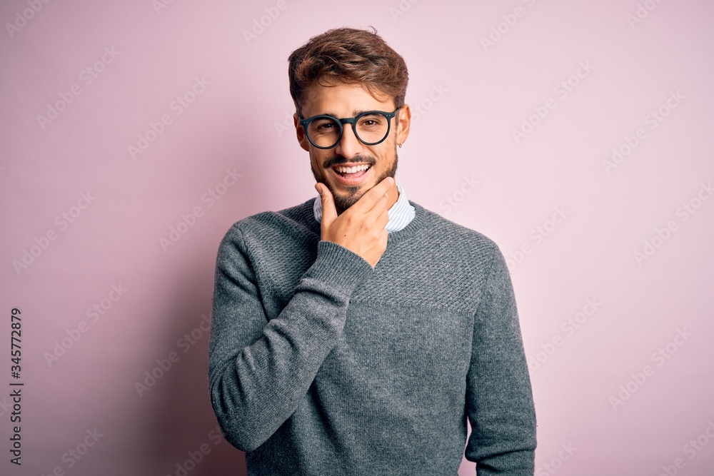 Young handsome man with beard wearing glasses and sweater standing over pink background looking confident at the camera smiling with crossed arms and hand raised on chin. Thinking positive. Young handsome man with beard wearing glasses and sweater standing over pink background looking confident at the camera smiling with crossed arms and hand raised on chin. Thinking positive.