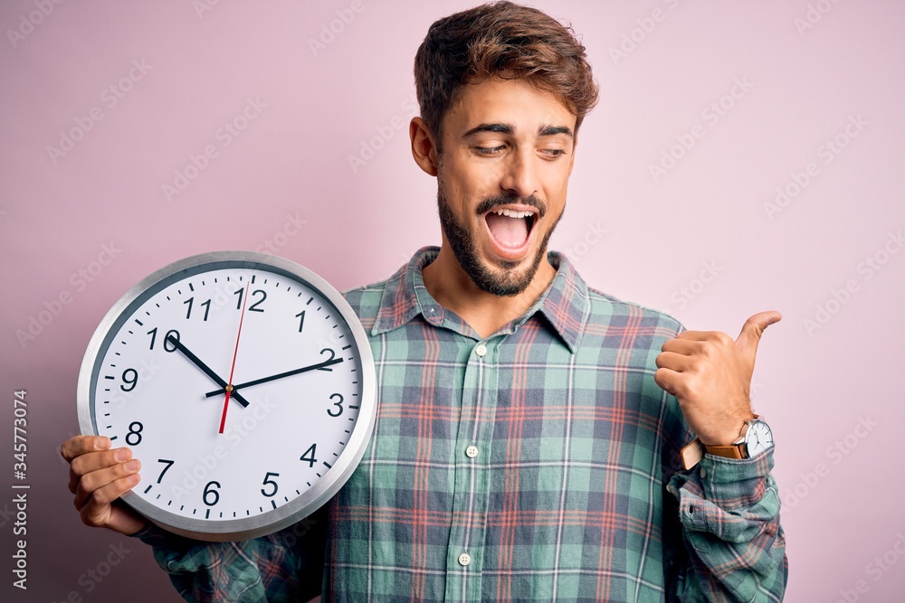 Young man with beard doing countdown using big clock over isolated pink background pointing and showing with thumb up to the side with happy face smiling Young man with beard doing countdown using big clock over isolated pink background pointing and showing with thumb up to the side with happy face smiling