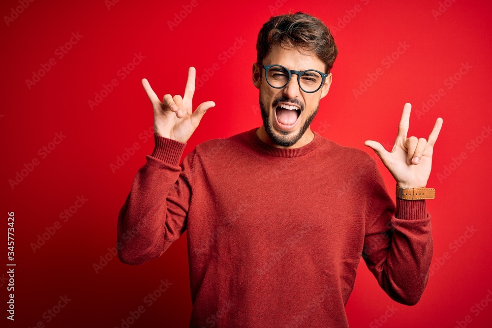 Young handsome man with beard wearing glasses and sweater standing over red background shouting with crazy expression doing rock symbol with hands up. Music star. Heavy music concept. Young handsome man with beard wearing glasses and sweater standing over red background shouting with crazy expression doing rock symbol with hands up. Music star. Heavy music concept.