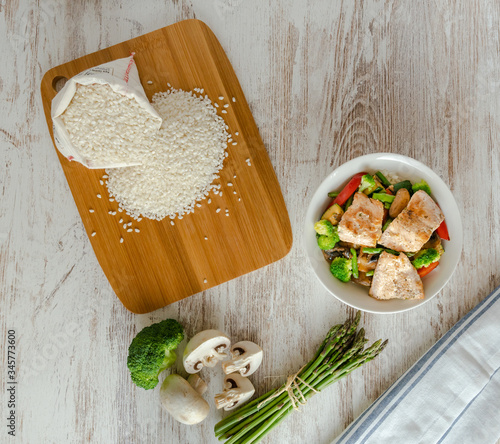 Cenital de bol de salmón salvaje, arroz, esparragos trigueros, brócoli, pimiento rojo, champiñones y saquito de arroz sobre mesa blanco. 