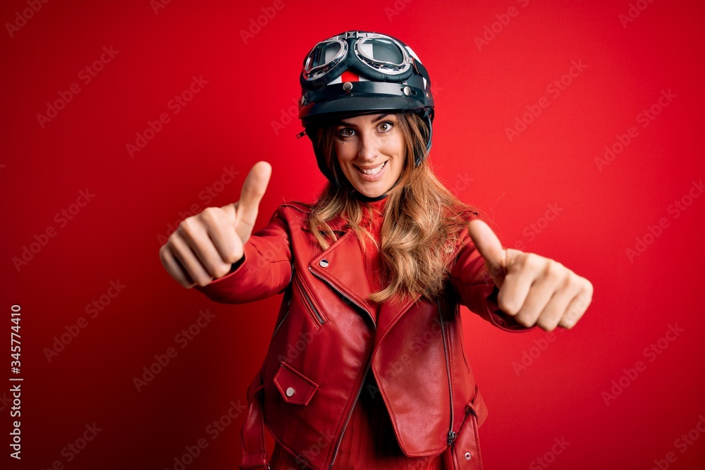 Young beautiful brunette motrocyclist woman wearing moto helmet over red background approving doing positive gesture with hand, thumbs up smiling and happy for success. Winner gesture. Young beautiful brunette motrocyclist woman wearing moto helmet over red background approving doing positive gesture with hand, thumbs up smiling and happy for success. Winner gesture.