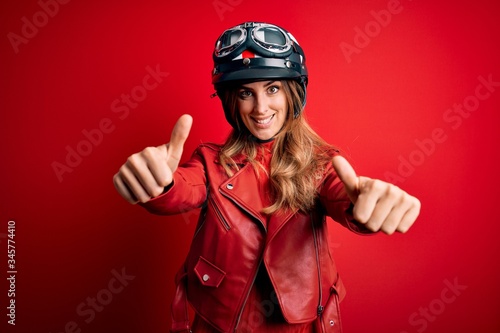 Young beautiful brunette motrocyclist woman wearing moto helmet over red background approving doing positive gesture with hand, thumbs up smiling and happy for success. Winner gesture.