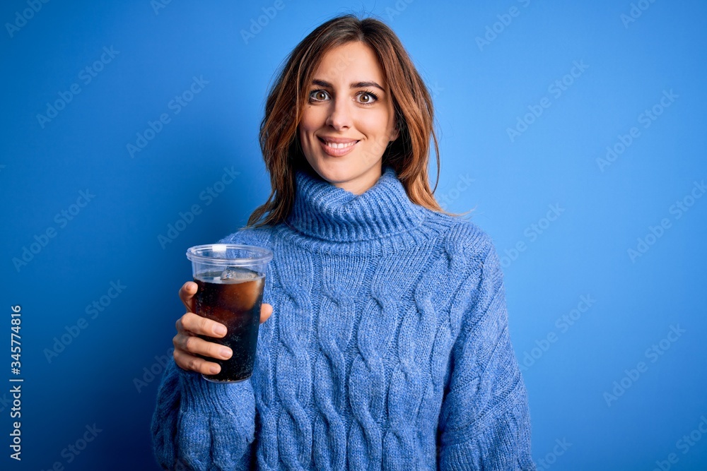 Young beautiful brunette woman drinking glass with cola refreshment over blue background with a happy face standing and smiling with a confident smile showing teeth Young beautiful brunette woman drinking glass with cola refreshment over blue background with a happy face standing and smiling with a confident smile showing teeth