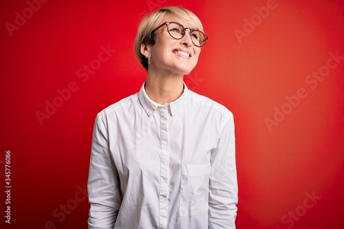 Young blonde business woman with short hair wearing glasses over red background looking away to side with smile on face, natural expression. Laughing confident.