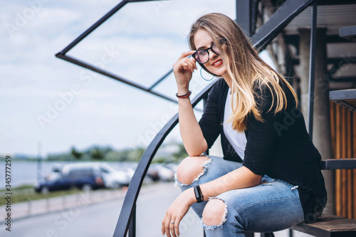 Beautiful girl with long hair and glasses sitting on metal stairs on the wooden background of house with vertical boards. Woman smiling and looking at camera