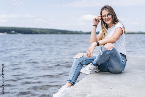 Young woman with long hair in stylish glasses posing on the concrete shore near the lake. Girl dressed in jeans and t-shirt smiling and looking at the camera