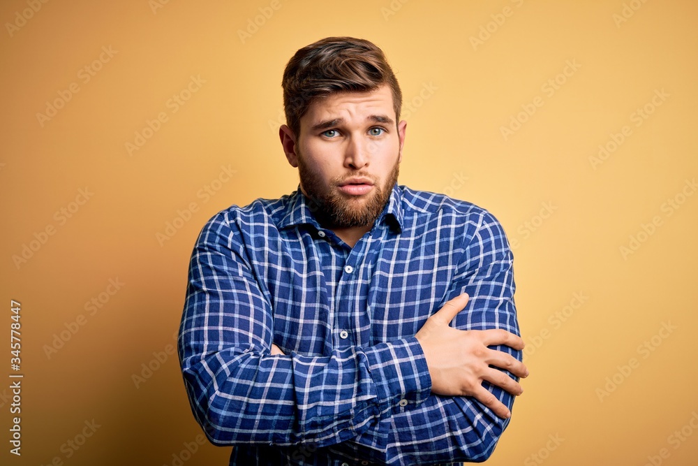 Young blond businessman with beard and blue eyes wearing shirt over yellow background shaking and freezing for winter cold with sad and shock expression on face Young blond businessman with beard and blue eyes wearing shirt over yellow background shaking and freezing for winter cold with sad and shock expression on face