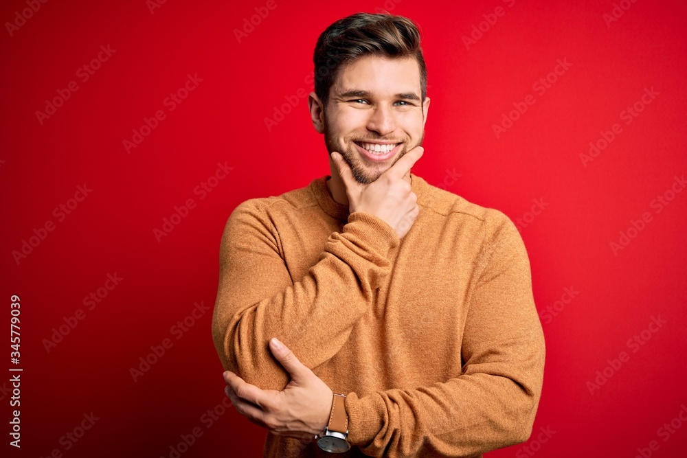 Young blond man with beard and blue eyes wearing casual sweater over red background looking confident at the camera smiling with crossed arms and hand raised on chin. Thinking positive.