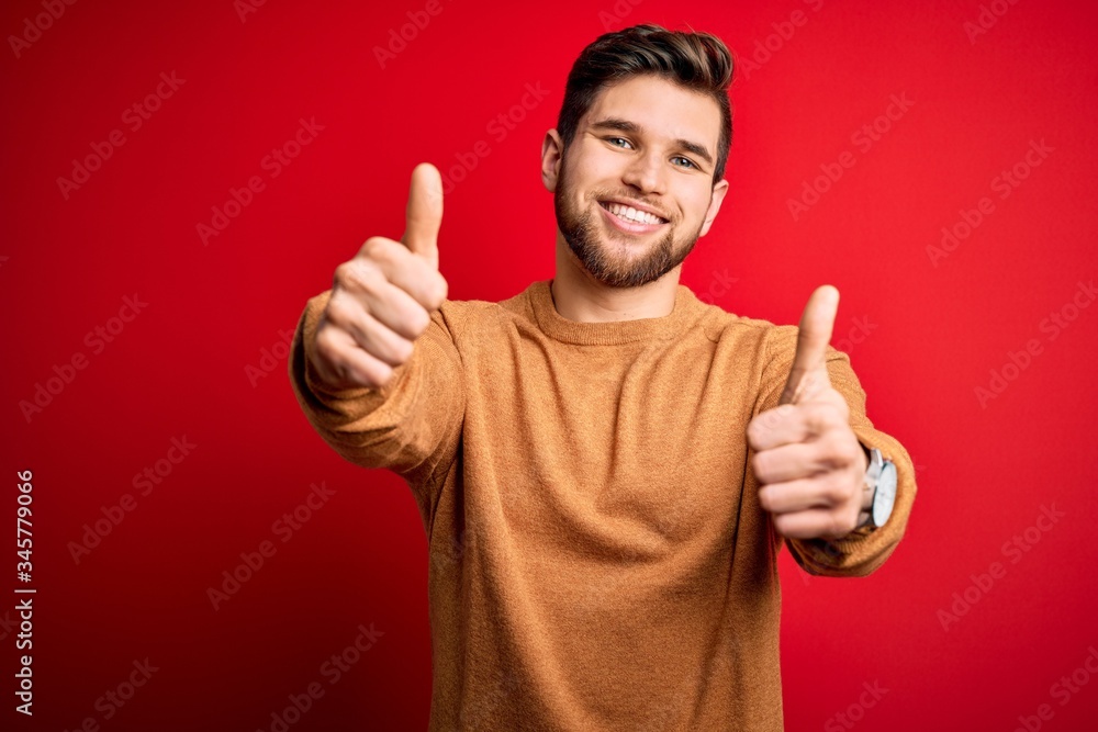 Young blond man with beard and blue eyes wearing casual sweater over red background approving doing positive gesture with hand, thumbs up smiling and happy for success. Winner gesture. Young blond man with beard and blue eyes wearing casual sweater over red background approving doing positive gesture with hand, thumbs up smiling and happy for success. Winner gesture.