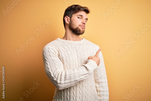 Young blond man with beard and blue eyes wearing white sweater over yellow background Pointing with hand finger to the side showing advertisement, serious and calm face