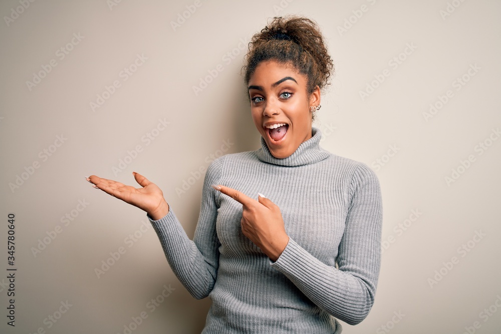 Beautiful african american girl wearing turtleneck sweater standing over white background amazed and smiling to the camera while presenting with hand and pointing with finger. Beautiful african american girl wearing turtleneck sweater standing over white background amazed and smiling to the camera while presenting with hand and pointing with finger.