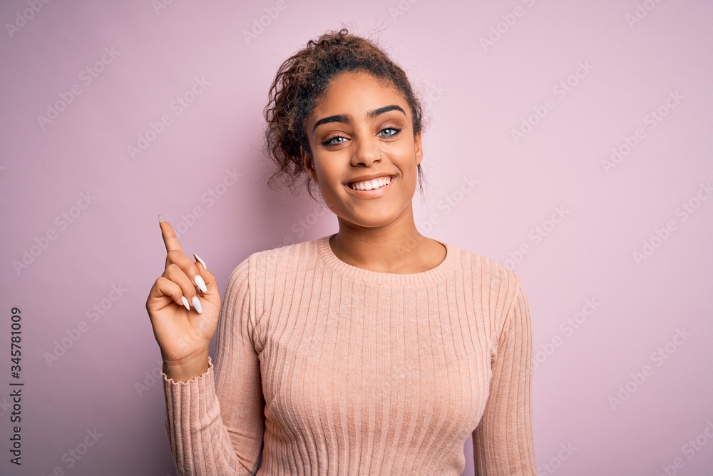 Young beautiful african american girl wearing casual sweater standing over pink background with a big smile on face, pointing with hand finger to the side looking at the camera. Young beautiful african american girl wearing casual sweater standing over pink background with a big smile on face, pointing with hand finger to the side looking at the camera.