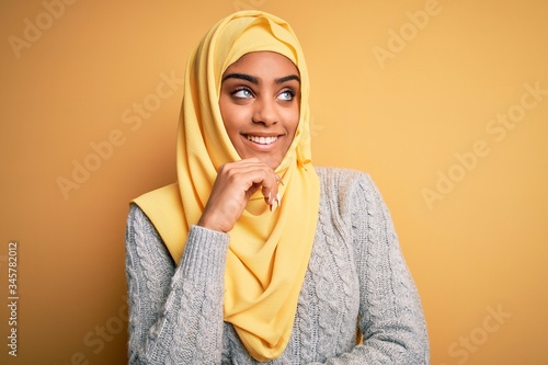 Young beautiful african american girl wearing muslim hijab over isolated yellow background with hand on chin thinking about question, pensive expression. Smiling and thoughtful face. Doubt concept. © Krakenimages.com