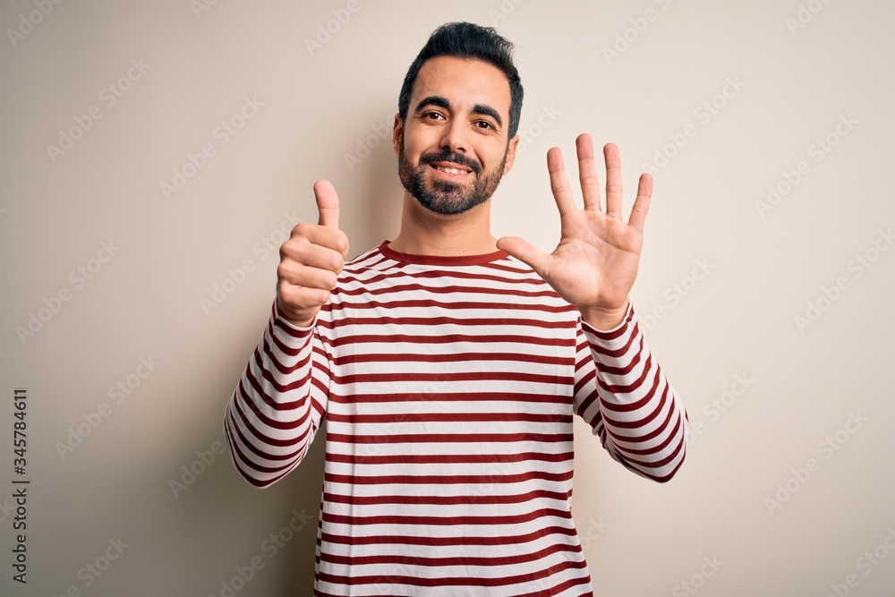 Fototapeta premium Young handsome man with beard wearing casual striped t-shirt standing over white background showing and pointing up with fingers number six while smiling confident and happy.