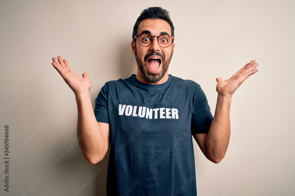 Handsome man with beard wearing t-shirt with volunteer message over white background celebrating crazy and amazed for success with arms raised and open eyes screaming excited. Winner concept Handsome man with beard wearing t-shirt with volunteer message over white background celebrating crazy and amazed for success with arms raised and open eyes screaming excited. Winner concept
